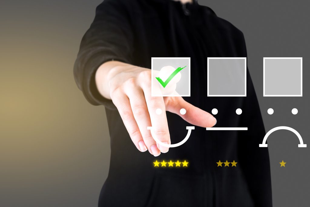 Uma pessoa vestindo roupa escura estende a mão e seleciona digitalmente uma avaliação positiva em uma interface interativa. O dedo toca um ícone de rosto sorridente acompanhado de um check verde e cinco estrelas, indicando alta satisfação. Ao lado, há outras opções de avaliação com expressões neutra e triste, cada uma com diferentes quantidades de estrelas. O fundo é escuro e desfocado, dando um efeito tecnológico e moderno, sugerindo um conceito de experiência do cliente e feedback digital.