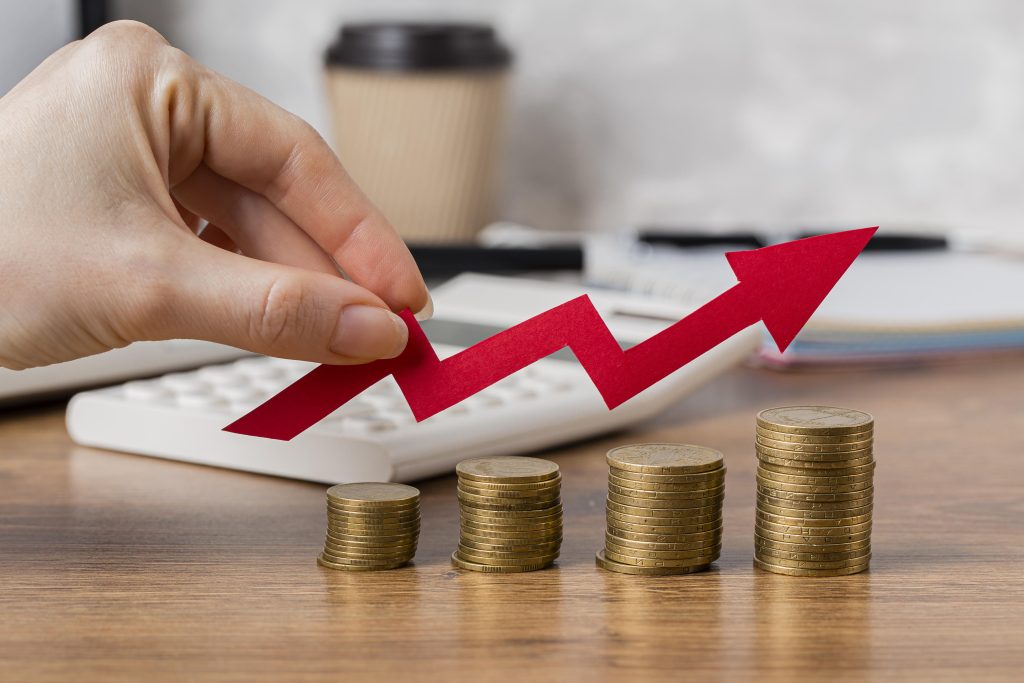 Uma mão segura uma seta vermelha em formato ascendente sobre pilhas crescentes de moedas douradas, simbolizando crescimento financeiro e investimentos bem-sucedidos. Ao fundo, há uma calculadora, um copo de café descartável e alguns cadernos sobre uma mesa de madeira, sugerindo um ambiente de planejamento financeiro. A imagem transmite a ideia de aumento de lucros, sucesso econômico e estratégias para a prosperidade financeira.