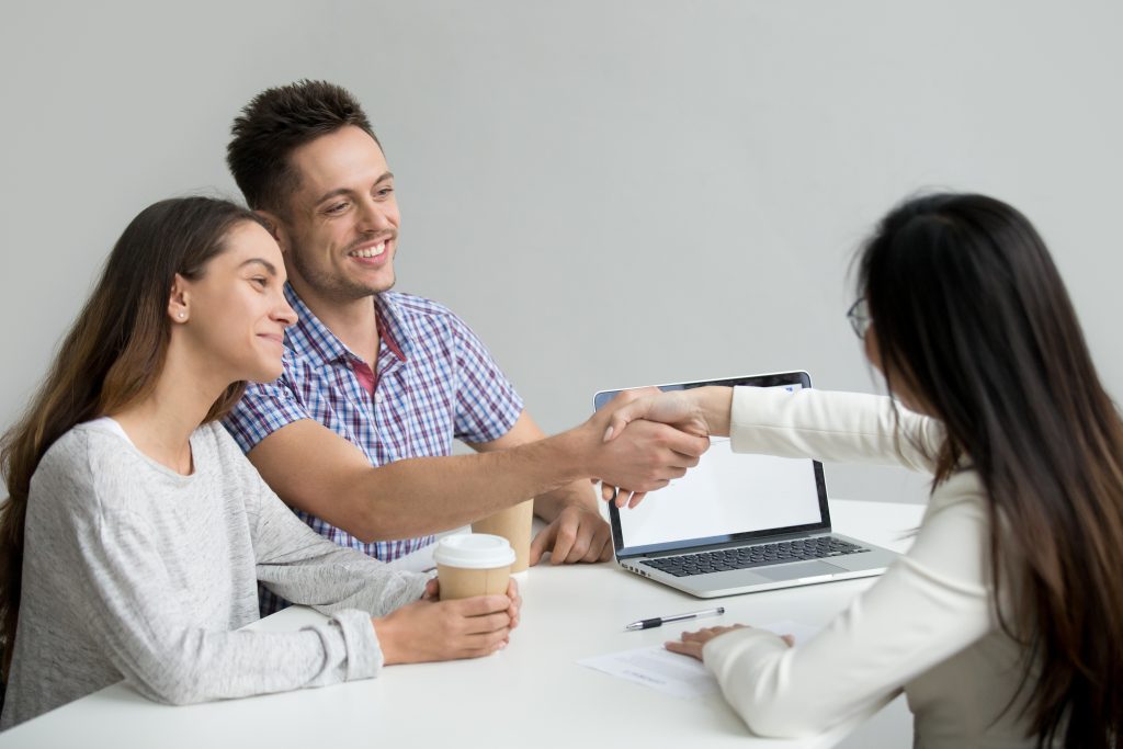 Um casal jovem e sorridente está reunido com uma consultora financeira em um ambiente profissional. O homem aperta a mão da consultora, simbolizando um acordo ou negociação bem-sucedida, enquanto a mulher segura um copo de café e observa com satisfação. Na mesa, há um laptop, documentos e uma caneta, reforçando a ideia de um compromisso financeiro ou imobiliário sendo firmado.