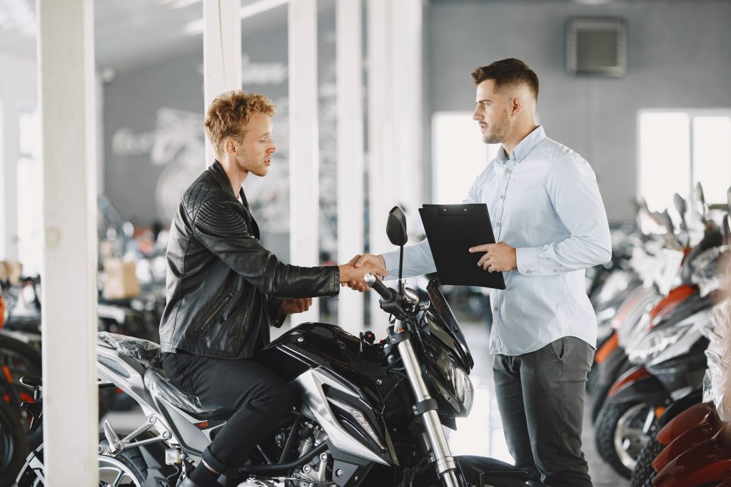 Um homem sentado em uma motocicleta preta aperta a mão de um vendedor em uma concessionária de motos. O cliente, vestindo uma jaqueta de couro preta, parece satisfeito com a negociação, enquanto o vendedor, segurando uma prancheta, confirma o acordo. O ambiente é repleto de motocicletas, indicando a compra ou financiamento de um novo veículo.