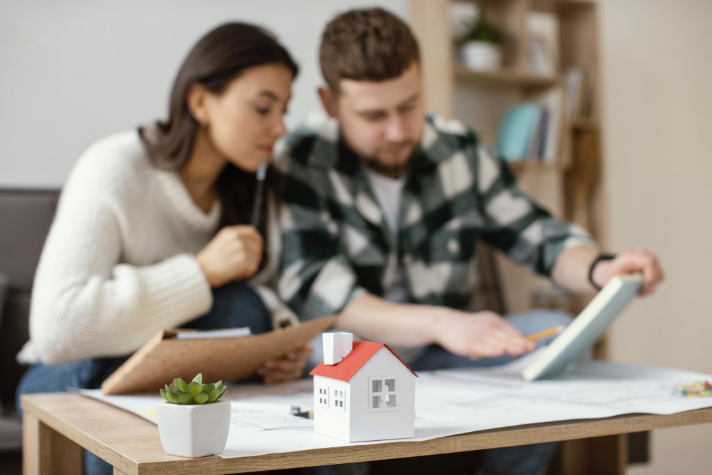 Um casal jovem analisa documentos financeiros e um livro enquanto planeja a compra de uma casa. No primeiro plano, há uma maquete de uma casa em miniatura com telhado vermelho sobre a mesa, simbolizando o objetivo da aquisição imobiliária. O ambiente é aconchegante, com uma decoração moderna e uma pequena planta sobre a mesa, reforçando a atmosfera de planejamento e futuro lar.