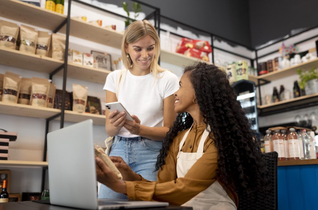 Uma vendedora sorridente, vestindo um avental bege e blusa marrom, está sentada atrás de um balcão em uma loja de produtos naturais, segurando um pacote de alimento e conversando com uma cliente. A cliente, uma jovem loira de camiseta branca e jeans, segura um smartphone e sorri enquanto interage com a vendedora. Ao fundo, prateleiras de madeira exibem diversos produtos embalados, bebidas e itens decorativos, criando um ambiente acolhedor e moderno. A imagem transmite um momento de atendimento atencioso e satisfação na experiência de compra.