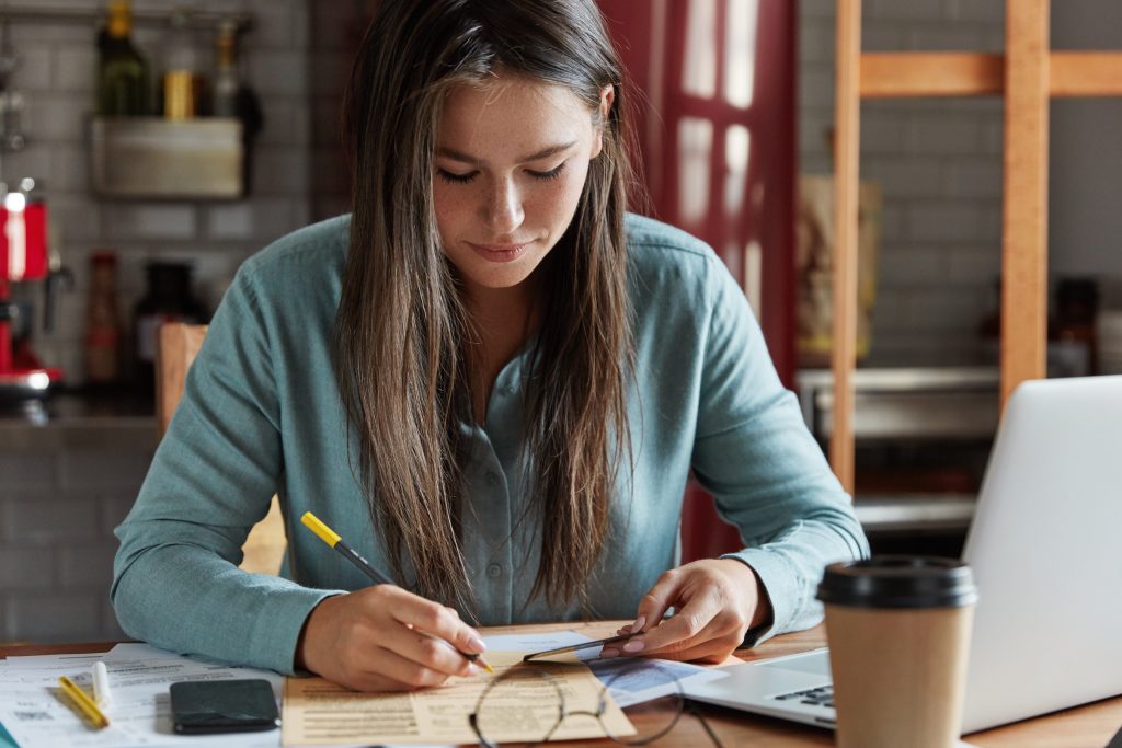Uma jovem concentrada escreve em um documento enquanto segura um cartão em um ambiente doméstico ou de escritório. Sobre a mesa, há um laptop, um smartphone, óculos, papéis e um copo de café, sugerindo que ela está trabalhando ou organizando informações financeiras ou profissionais. O cenário de fundo traz uma cozinha com itens decorativos, reforçando a ideia de home office.