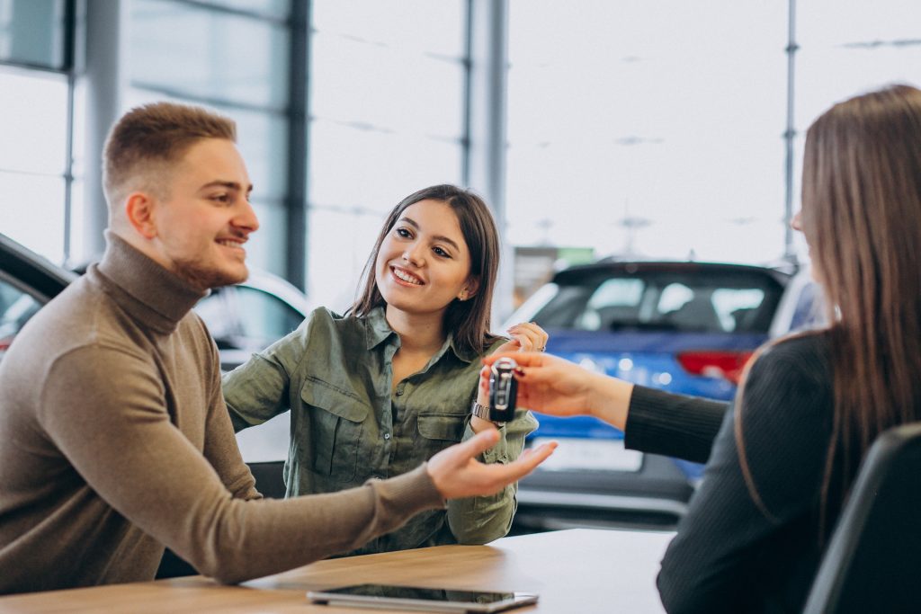 Um jovem casal está em uma concessionária de carros, sorrindo enquanto recebe as chaves de um veículo de uma vendedora. O homem, vestindo um suéter bege, estende a mão para pegar as chaves, enquanto a mulher, com uma camisa verde, observa a cena com entusiasmo. Ao fundo, há carros expostos e amplas janelas, criando um ambiente moderno e iluminado. A imagem transmite satisfação e a emoção da compra de um novo carro.