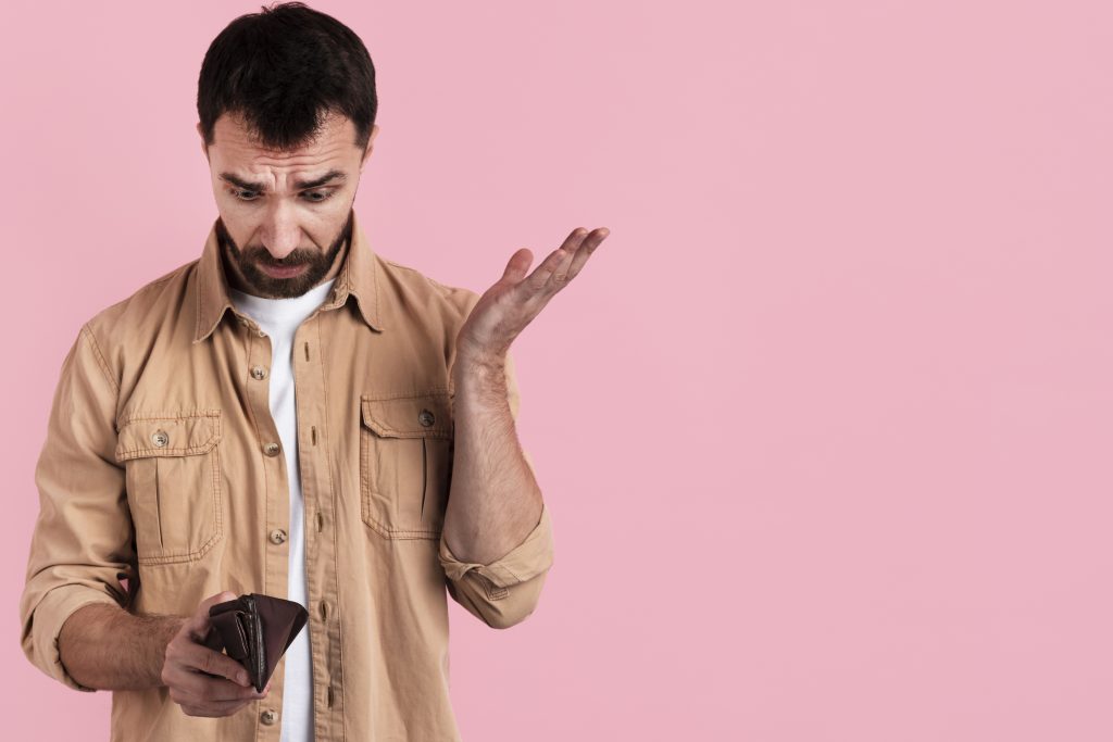 Um homem de expressão preocupada olha para sua carteira vazia enquanto levanta a outra mão em um gesto de frustração. Ele veste uma jaqueta bege sobre uma camiseta branca e está diante de um fundo rosa sólido, transmitindo uma sensação de surpresa e inquietação financeira.