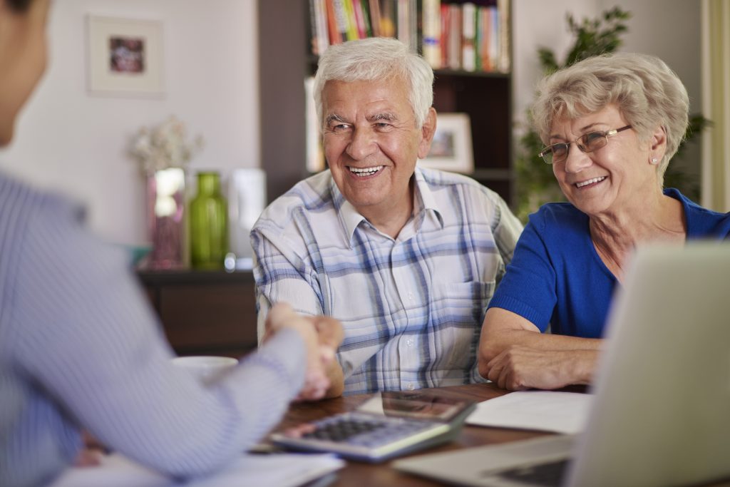 Um casal de idosos sorridente está sentado em uma mesa, conversando com uma profissional. O homem, vestindo uma camisa xadrez, aperta a mão da consultora, enquanto a mulher, de blusa azul e óculos, observa a interação com um sorriso. Na mesa, há um laptop, uma calculadora e documentos, sugerindo uma reunião sobre finanças ou planejamento. O ambiente é aconchegante, com uma estante de livros e elementos decorativos ao fundo, transmitindo uma atmosfera de confiança e satisfação.