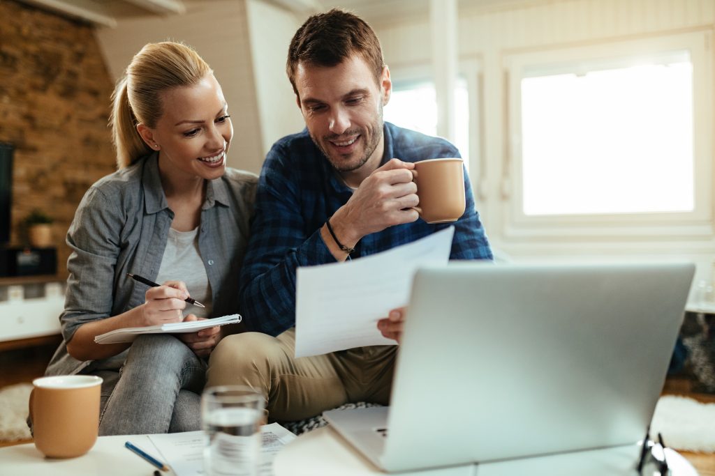 Um casal sorridente está sentado em casa analisando documentos financeiros enquanto usa um laptop. O homem, vestindo uma camisa xadrez azul, segura uma caneca de café e observa os papéis com atenção. A mulher, vestindo uma camisa cinza, segura um caderno e uma caneta, parecendo anotar informações enquanto olha para o parceiro. A cena transmite um ambiente aconchegante e colaborativo, sugerindo planejamento financeiro ou organização do orçamento doméstico.
