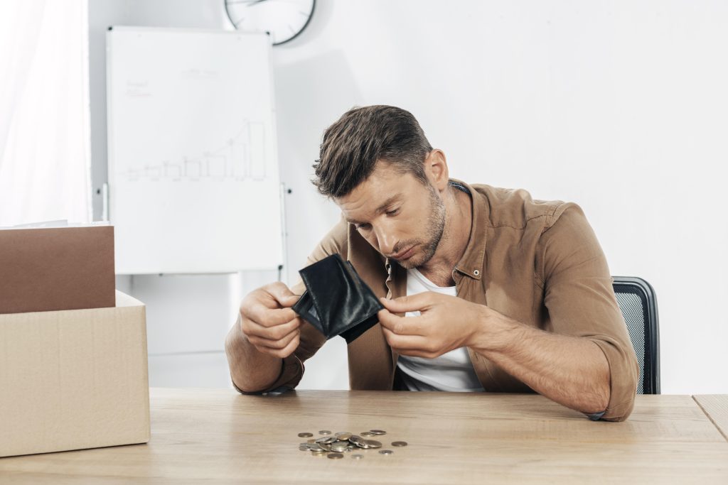 Um homem com expressão preocupada está sentado à mesa, segurando uma carteira vazia e olhando para algumas moedas espalhadas na superfície. Ele veste uma camisa marrom e parece refletir sobre sua situação financeira. No fundo, há um quadro branco com gráficos desenhados e um relógio na parede, sugerindo um ambiente de escritório. Uma caixa de papelão ao lado indica um possível momento de mudança ou dificuldade financeira. A imagem transmite a sensação de preocupação com dinheiro e desafios econômicos.