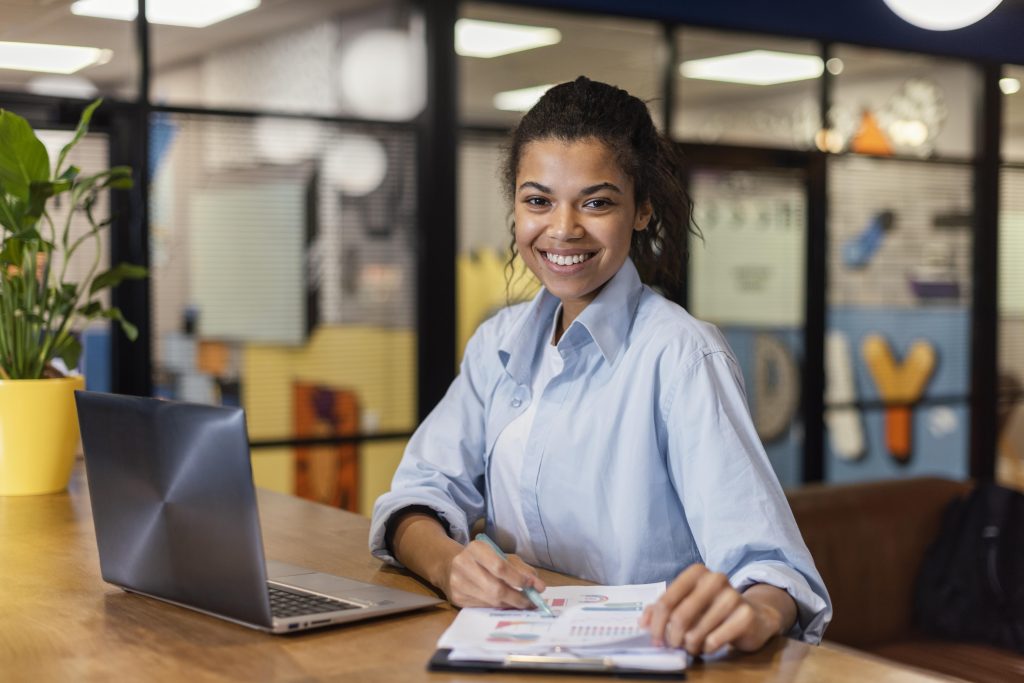 Uma jovem profissional sorridente está sentada em um ambiente de escritório moderno, analisando documentos com gráficos enquanto segura uma caneta. Ela veste uma camisa azul clara e transmite uma expressão de confiança e positividade. Sobre a mesa de madeira, há um laptop aberto e um vaso de planta, reforçando um ambiente produtivo e organizado. O fundo colorido e decorado sugere um espaço de trabalho criativo e colaborativo.