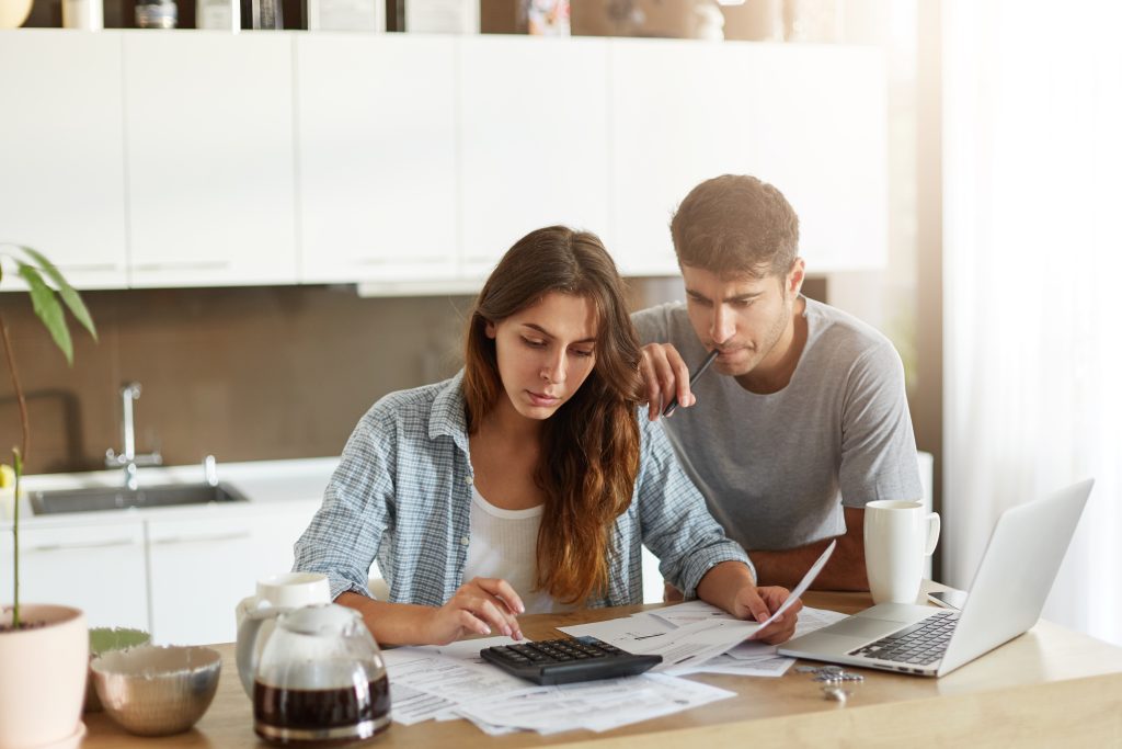 Um jovem casal está na cozinha analisando seu orçamento familiar. A mulher, vestindo uma camisa xadrez, usa uma calculadora enquanto segura um documento, demonstrando concentração. O homem, de camiseta cinza, observa atentamente com uma expressão séria, segurando uma caneta próxima ao rosto. Na mesa, há um laptop, papéis espalhados, uma cafeteira e xícaras, sugerindo um momento de planejamento financeiro e tomada de decisões importantes.