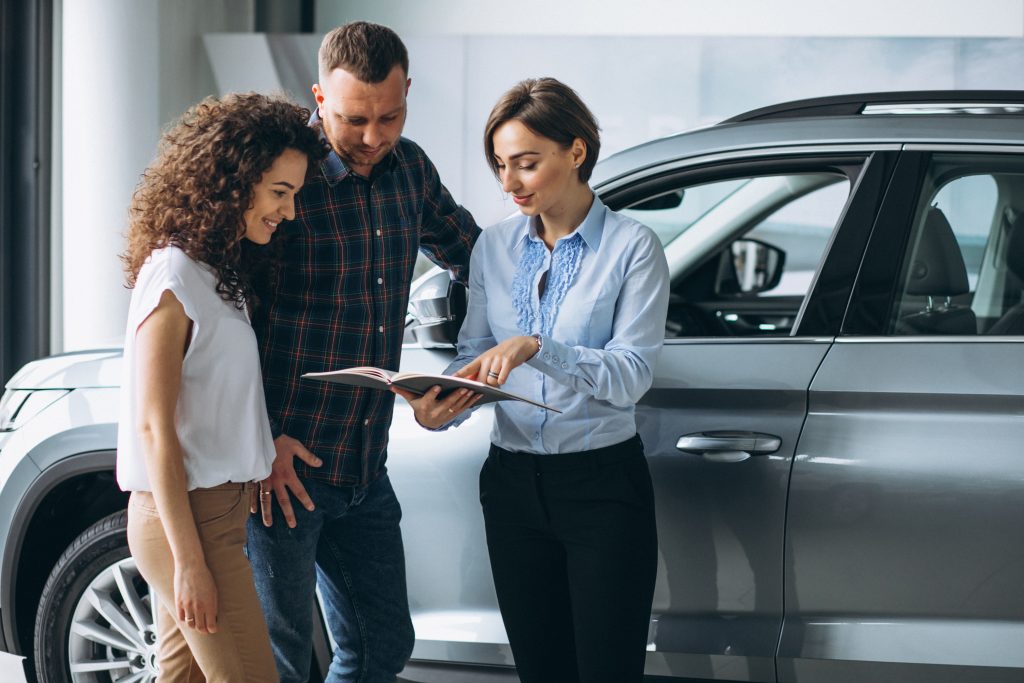 Um casal jovem conversa com uma vendedora em uma concessionária de carros. A vendedora, vestindo uma camisa azul, aponta para um catálogo enquanto explica detalhes sobre o veículo prateado ao lado deles. O casal, sorridente e atento, demonstra interesse, indicando que estão considerando a compra ou financiamento do carro.
